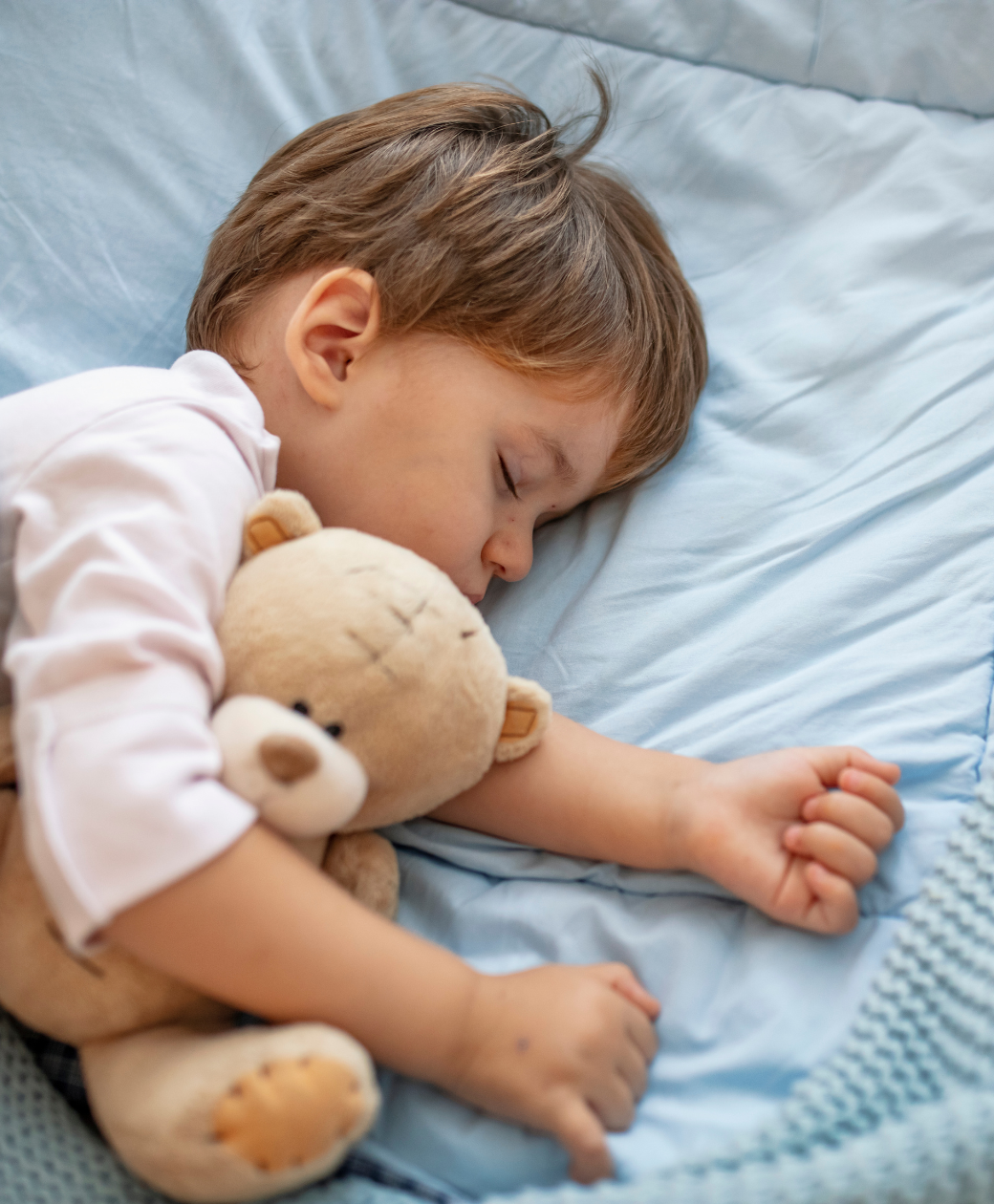 Toddler sleeping peacefully in bed while cuddling a teddy bear, illustrating night time potty training and staying dry overnight.